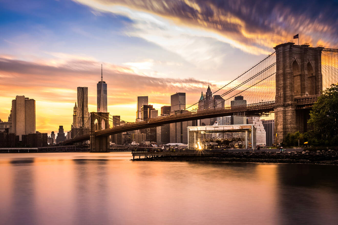 View of the Brooklyn Bridge and Manhattan skyline at sunset, with colorful clouds reflecting in the calm water of the East River.